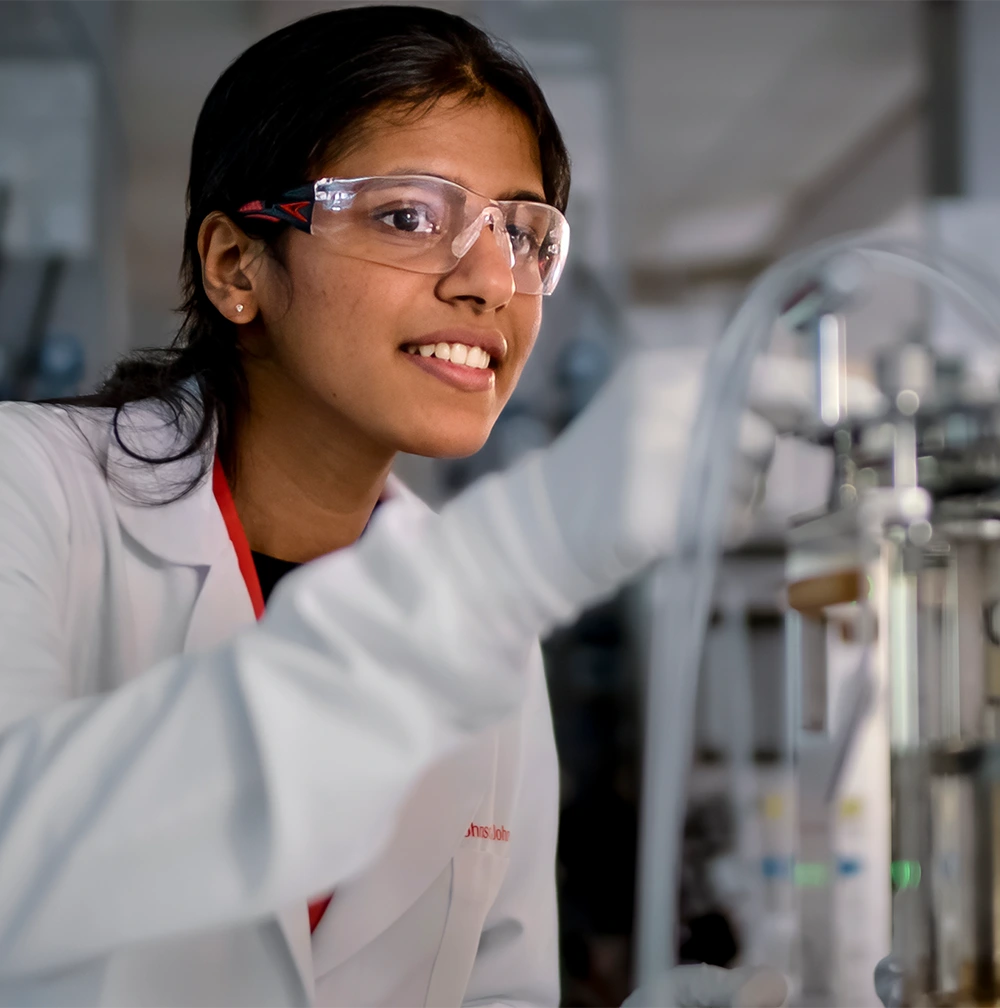 A young Johnson & Johnson associate wearing a lab coat, gloves, and safety googles works with a mini-bioreactor (photo)